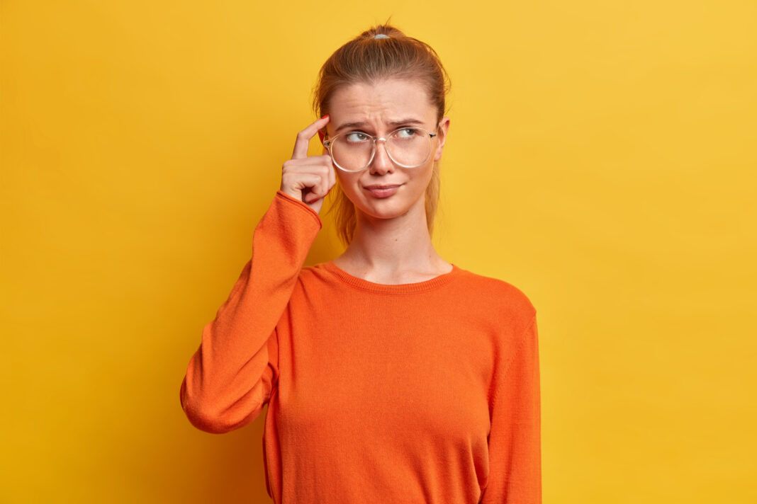 portrait-thoughtful-serious-woman-keeps-finger-temple-tries-focus-something-wears-casual-orange-jumper-poses0