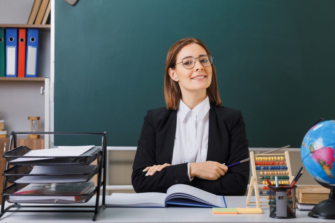 young woman teacher wearing glasses sitting at school desk in front of blackboard in classroom with abacus and globe checking class register holding pointer smiling confident0